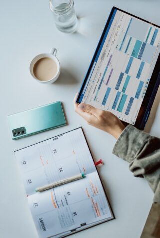 person with a tablet in their hands, displaying an online calendar with appointments, next to a cell phone, and a planner