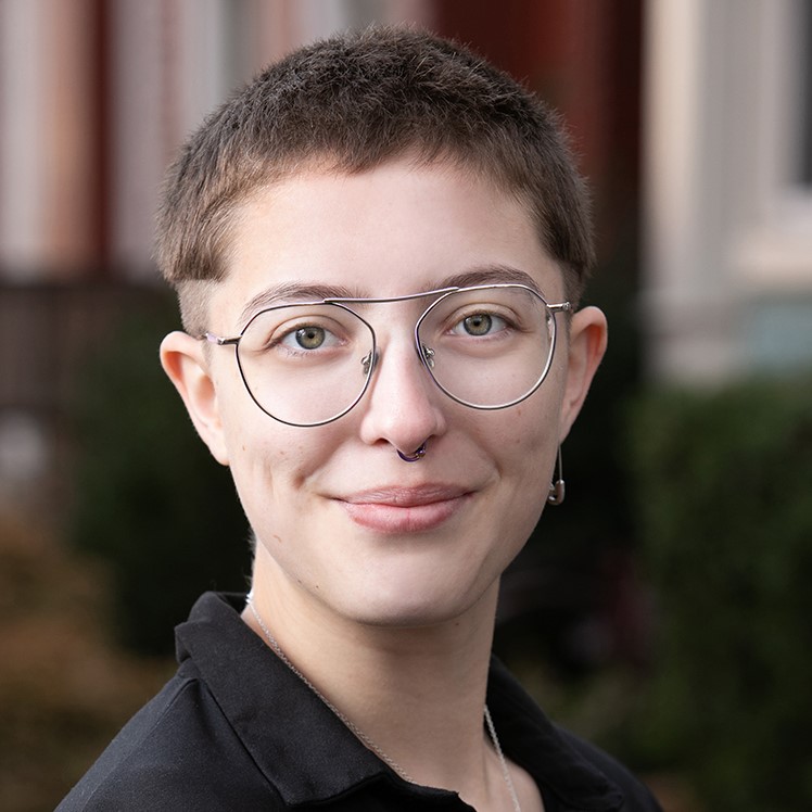 Head shot of Roz. Roz is smiling and standing outside in front of a city home. They are wearing glasses and a black shirt.