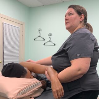 Kai works on her client's shoulder. The client is face-down and supported with pillows. Kai stands to the client's left just below the shoulder. She supports the client's arm while it is outstretched so that she can work beneath the shoulder blade.