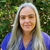 Head Shot of Veda. She has long silver hair and is wearing a purple blouse. The green leaves of a bush are behind her.