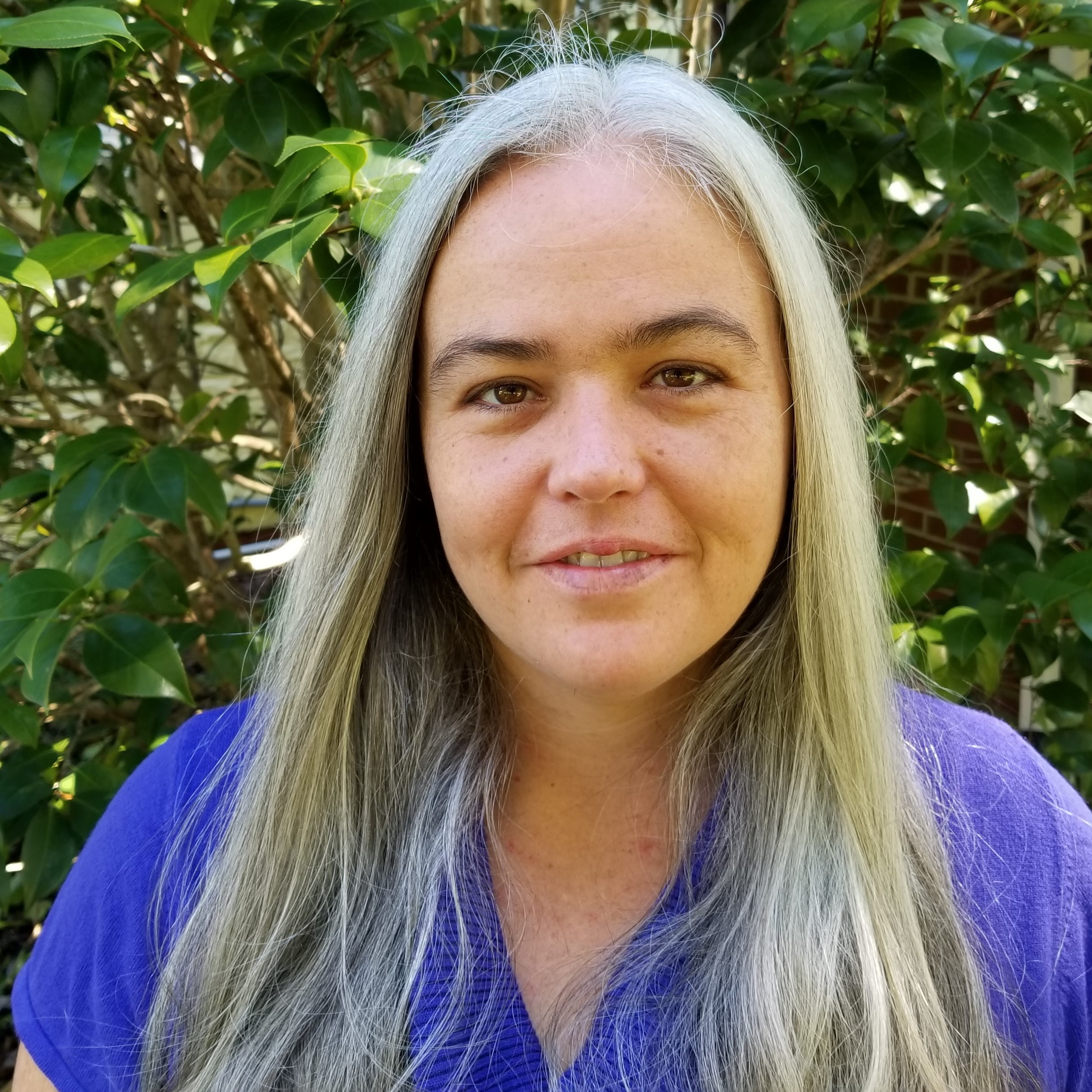 Head Shot of Veda. She has long silver hair and is wearing a purple blouse. The green leaves of a bush are behind her.